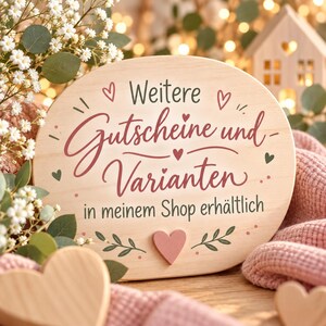 May include: A light-colored oval wooden sign featuring the German text "Weitere Gutscheine und Varianten in meinem Shop erh&auml;ltlich" in dark script and print. The sign is decorated with small painted hearts, leaves, and a central wooden heart.