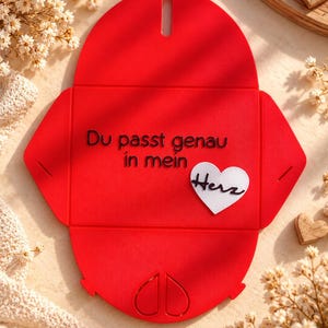 May include: A bright red, flat-packed envelope made of a textured material. The front features the black text 'Du passt genau in mein' and a white heart-shaped cutout with the black script word 'Herz' attached to the center.