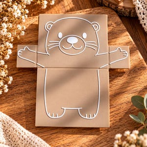 May include: A flat, light brown, rectangular card or paper cutout featuring a white line drawing of an otter with its arms outstretched. The otter has a rounded head, small ears, whiskers, and a simple body outline. The design is centered on the card.