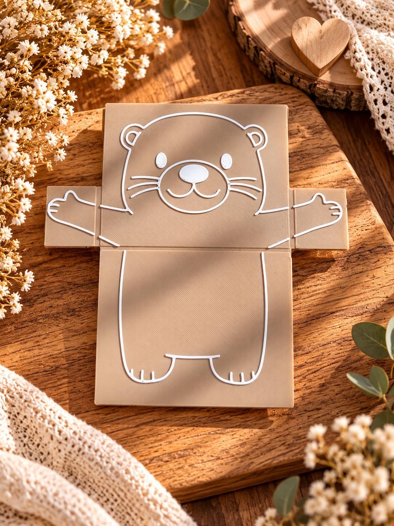 May include: A flat, light brown, rectangular card or paper cutout featuring a white line drawing of an otter with its arms outstretched. The otter has a rounded head, small ears, whiskers, and a simple body outline. The design is centered on the card.