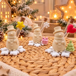 Peut inclure: Trois figurines de bonhomme de neige couleur crème, portant un haut-de-forme et une écharpe, posées sur des socles en forme de flocon blanc. Chaque bonhomme tient un tube à essai en verre avec un bouchon en liège, une étoile, un flocon ou un cœur.