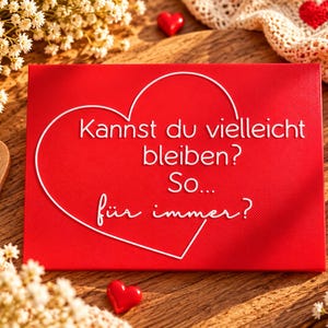 May include: A rectangular red sign featuring a white outline of a heart and the German text: Kannst du vielleicht bleiben? So... f&uuml;r immer? The sign is surrounded by small white flowers, wooden heart cutouts, and red heart accents.