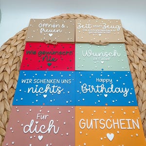 Peut inclure: Huit cartes rectangulaires de diff&eacute;rentes couleurs, notamment beige, rouge, vert clair, bleu et orange. Chaque carte pr&eacute;sente du texte blanc et de petits points blancs. Le texte inclut des phrases comme '&Ouml;ffnen & freuen', 'Zeit statt Zeug', 'Wunsch erf&uuml;ller', 'Happy Birthday' et 'GUTSCHEIN'.