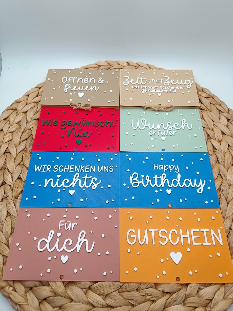 Peut inclure: Huit cartes rectangulaires de diff&eacute;rentes couleurs, notamment beige, rouge, vert clair, bleu et orange. Chaque carte pr&eacute;sente du texte blanc et de petits points blancs. Le texte inclut des phrases comme '&Ouml;ffnen & freuen', 'Zeit statt Zeug', 'Wunsch erf&uuml;ller', 'Happy Birthday' et 'GUTSCHEIN'.