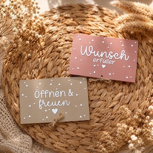 May include: Two rectangular cards with white text and dots. The top card is dusty rose with the text "Wunsch erfüller" and a heart. The bottom card is tan with the text "Öffnen & freuen" and a heart. Both cards feature small twine accents.