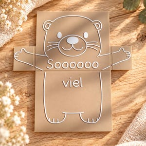 May include: A tan rectangular plaque featuring a white line art illustration of a bear with outstretched arms. The text "Soooooo" is printed above the bear's midsection, and the word "viel" is printed below it in white lettering.