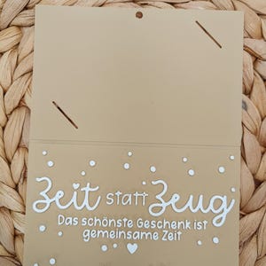 Peut inclure: Une carte rectangulaire beige avec un pli en haut, pr&eacute;sentant le texte blanc "Zeit statt Zeug" et "Das sch&ouml;nste Geschenk ist gemeinsame Zeit" imprim&eacute; sur la moiti&eacute; inf&eacute;rieure, agr&eacute;ment&eacute; de petits points blancs et d'un c&oelig;ur.