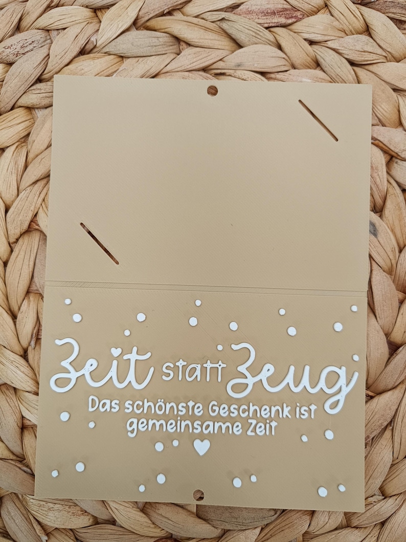 Peut inclure: Une carte rectangulaire beige avec un pli en haut, pr&eacute;sentant le texte blanc "Zeit statt Zeug" et "Das sch&ouml;nste Geschenk ist gemeinsame Zeit" imprim&eacute; sur la moiti&eacute; inf&eacute;rieure, agr&eacute;ment&eacute; de petits points blancs et d'un c&oelig;ur.