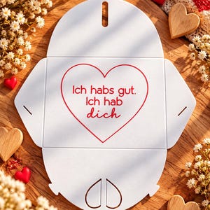 May include: A white flat-pack paper envelope with a die-cut heart shape in the center. The red text inside the heart reads, "Ich habs gut. Ich hab dich." The envelope features rounded flaps and a small heart-shaped cutout at the bottom.