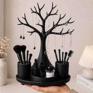 May include: A matte black jewellery and cosmetic organiser featuring a central tree-shaped stand with multiple branches for hanging necklaces and earrings. The base includes two cylindrical brush holders and a central tray holding small bottles and containers.