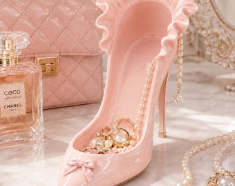 May include: A glossy light pink high-heeled shoe figurine featuring a ruffled collar, a small bow on the toe, and a dangling pearl chain. The interior of the shoe holds a collection of gold-toned rings and pearl jewelry pieces.