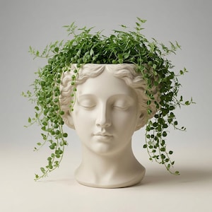 Op de afbeelding: Een matte gebroken witte keramische plantenbak in de vorm van een menselijk hoofd met gesloten ogen en golvend haar. De bovenkant van het hoofd is open en bevat een weelderige, hangende groene plant met kleine ronde blaadjes aan de zijkanten.