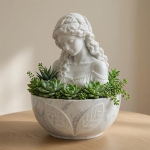 Puede incluir: Una maceta de mármol blanco con el busto esculpido de una persona de cabello largo y ondulado. La base en forma de cuenco está decorada con patrones de hojas talladas y llena de una variedad de plantas suculentas verdes, incluyendo rosetas.