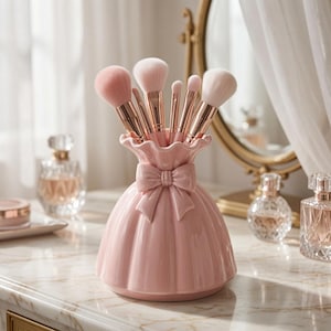 Pode incluir: Um suporte de cerâmica para pincéis de maquilhagem em rosa claro com formato de saia rodada e um laço decorativo no pescoço. O suporte contém vários pincéis com virolas em ouro rosa e cerdas claras, dispostos na vertical.