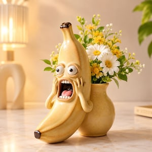May include: A yellow ceramic vase shaped like a banana with a wide-eyed, screaming facial expression and hands pressed against its cheeks. The banana figure is attached to a rounded, speckled yellow vase containing a bouquet of white and yellow flowers.