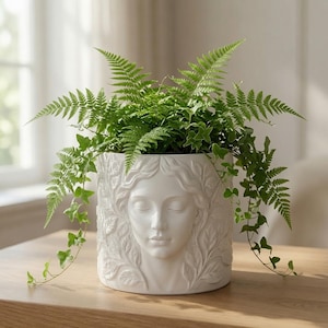 May include: A white ceramic cylindrical planter featuring a relief sculpture of a serene human face surrounded by detailed foliage. A lush green fern and trailing ivy plant are potted inside, with leaves extending over the top rim of the vessel.