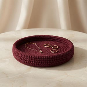 May include: A round, deep-walled tray crafted from dark burgundy coiled rope. Inside the tray, there is a delicate gold chain necklace and three gold rings, including one ring featuring a small, clear, round gemstone set in the center.