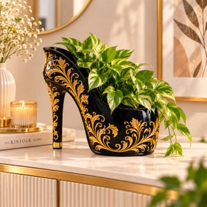 May include: A black ceramic planter shaped like a high-heeled shoe, featuring intricate gold filigree scrollwork patterns along the exterior. The planter holds a lush green and yellow variegated pothos plant with trailing vines.