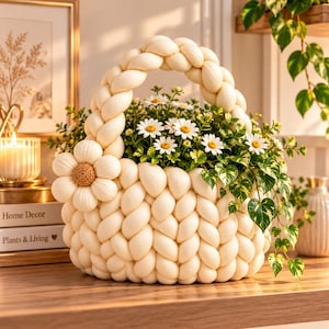 Könnte beinhalten: Ein cremefarbener, grob gestrickter Korb mit geflochtenem Henkel und einer großen, plastischen Blumenapplikation an der Seite. Der Korb ist mit einem üppigen Arrangement aus weißen Gänseblümchen und grün-weiß panaschierten Efeublättern gefüllt.