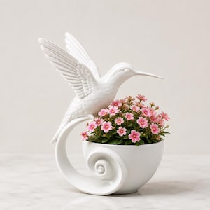 Può includere: Un vaso in ceramica bianca raffigurante un colibrì scolpito con le ali spiegate verso l'alto, appoggiato su una base curva a spirale. Il vaso contiene un gruppo di piccoli fiori rosa vivaci con foglie verdi, creando un display decorativo.