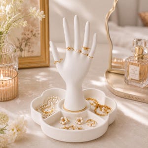 May include: A white ceramic hand-shaped jewellery display stand featuring a flower-shaped base with multiple compartments. The stand holds several gold-toned rings on its fingers, while the base contains gold-toned necklaces, bracelets, and floral-shaped rings.