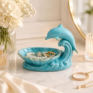 Könnte beinhalten: Eine glänzende türkisfarbene Schmuckschale aus Keramik in Form einer Muschel, die einen aus einer Welle springenden Delfin darstellt. Die Ränder der Muschel und die Flossen des Delfins sind mit einem dünnen Goldrand verziert. Sie enthält ein Perlenarmband.