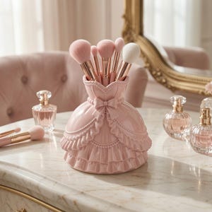 Op de afbeelding: Een lichtroze keramische make-up kwastenhouder in de vorm van een baljurk met ruches en een strik in het midden. De houder bevat vijf make-up kwasten met roségouden hulzen en roze of witte haren, staand op een marmeren oppervlak.