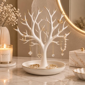 May include: A white jewelry stand shaped like a tree with gold-tipped branches and a circular base. The stand holds several gold necklaces and rings, with additional rings resting in the tray at the base. The design features smooth, matte white surfaces with gold accents on the branch tips and the rim of the tray.