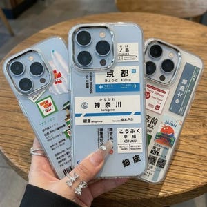 以下が含まれることがあります： 駅の看板、地下鉄のチケット、コンビニのロゴ、日本語の文字など、日本をテーマにしたグラフィックデザインが背面にプリントされた、3つの透明なスマートフォンケース。それぞれの保護カバーには、詳細な日本の風景や標識が描かれています。