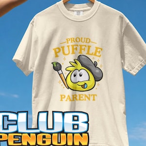 May include: An ivory short-sleeve t-shirt featuring a graphic of a yellow Puffle character wearing a grey beret and holding a paintbrush. The text reads PROUD PUFFLE PARENT and CLUB PENGUIN. The shirt is displayed on a wooden hanger against a blue sky.