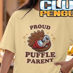 May include: A light yellow short-sleeve t-shirt featuring a brown cartoon puffle character with large eyes and red goggles. The text above the character reads PROUD and below it reads PUFFLE PARENT, with small star accents surrounding the graphic.