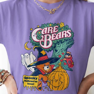 May include: A violet t-shirt featuring a graphic print of Care Bears characters in a Halloween scene. The design includes a bear in a witch hat carving a pumpkin, a ghost, a tree, and the text Care Bears and Spooky Halloween issue.