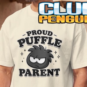 May include: An ivory short-sleeve t-shirt featuring a graphic of a black Puffle character with a grumpy expression. The text reads "PROUD PUFFLE PARENT" in black letters, surrounded by small star icons. The shirt has a crew neckline.