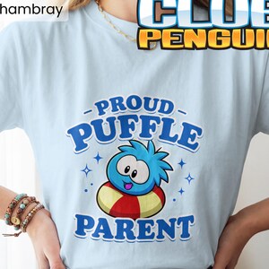 May include: A light blue t-shirt featuring a graphic of a blue Puffle character inside a red and yellow striped inner tube. The text reads PROUD PUFFLE PARENT in blue letters, with small star accents surrounding the character. The shirt is labelled Chambray.