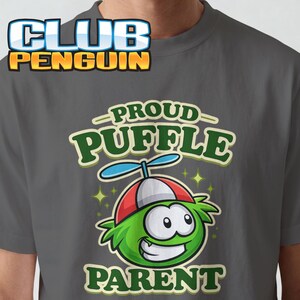 May include: A dark grey t-shirt featuring a graphic of a green, furry creature wearing a red and white propeller hat. The text CLUB PENGUIN appears at the top, while the text PROUD PUFFLE PARENT is printed in green below the creature illustration.