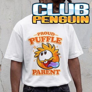 May include: A white t-shirt featuring a graphic of a round, orange, spiky creature with large eyes, a wide mouth, and a purple tongue sticking out. The text above the creature reads PROUD PUFFLE and the text below reads PARENT in orange letters.