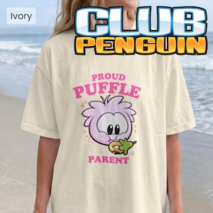 May include: An ivory t-shirt featuring a graphic of a light purple Puffle character eating a green snack. The text above reads PROUD PUFFLE and the text below reads PARENT in pink letters. Small pink star accents surround the character.