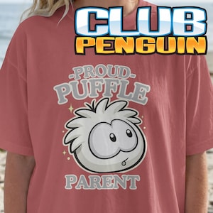 May include: A dusty rose t-shirt featuring a graphic of a white, fluffy Puffle character from Club Penguin. The text above the character reads PROUD and PUFFLE, while the text below reads PARENT. The design includes small star accents.