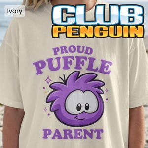 May include: An ivory short-sleeve t-shirt featuring a graphic of a purple Puffle character from Club Penguin. The text reads PROUD PUFFLE PARENT in purple letters above and below the character, with small star accents surrounding the graphic.