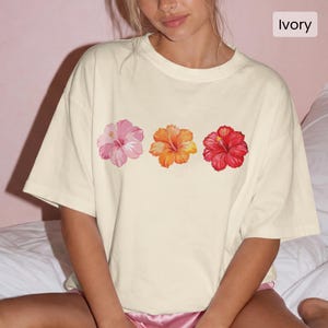 May include: An ivory-colored short-sleeve t-shirt featuring a horizontal graphic print of three hibiscus flowers in a row. The flowers are colored in shades of light pink, orange-yellow, and deep red. The text Ivory is printed in the corner.