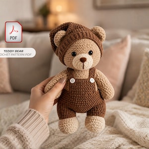 May include: A crochet teddy bear toy in light tan yarn, wearing dark brown overalls with two white buttons and a matching dark brown knit cap. The bear has black bead eyes and a dark brown stitched nose. Text reads TEDDY BEAR CROCHET PATTERN PDF.