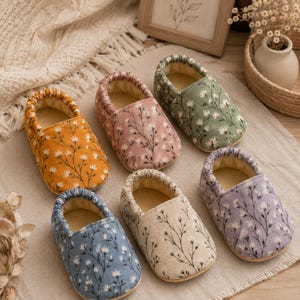 Puede incluir: Seis patucos de bebé suaves dispuestos en grupo, cada uno con una base de color sólido distinta que incluye amarillo mostaza, rosa empolvado, verde salvia, azul pizarra, crema y lavanda, decorados con un patrón de ramas florales negras.