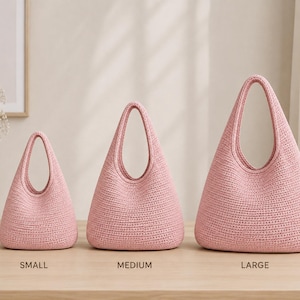 Könnte beinhalten: Drei rosa gehäkelte Handtaschen in einer Reihe, beschriftet mit small, medium und large. Jede Tasche hat einen einzelnen integrierten Griff, einen abgerundeten Boden und ein strukturiertes Webmuster. Die Taschen sind identisch im Design.