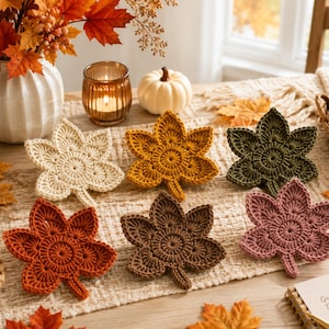 May include: A set of six crochet leaf coasters in cream, mustard yellow, olive green, burnt orange, dark brown, and dusty rose. Each coaster features a circular center with five radiating leaf points, crafted with detailed yarn stitching.