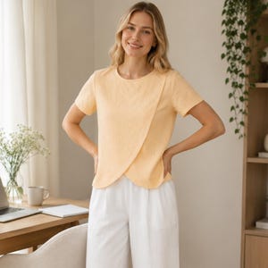 May include: A short-sleeved, light peach-colored top featuring a rounded neckline and a distinctive overlapping front panel design that creates a curved hemline, paired with loose-fitting, solid white trousers with a clean, simple aesthetic.