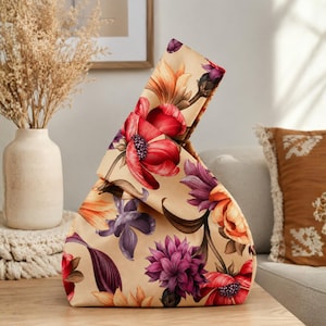 May include: A fabric knot bag featuring a beige base color with a vibrant floral pattern. The design includes large, detailed flowers in shades of red, orange, purple, and dark burgundy. The bag has a unique asymmetrical handle formed by the fabric.