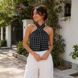 May include: A black and white plaid halter top featuring a high neckline with a crisscross design, paired with solid white trousers. The sleeveless top has a fitted silhouette and a classic checkered pattern throughout the fabric.