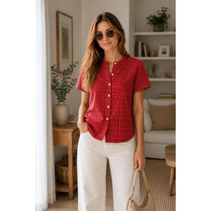 May include: A short-sleeved red button-down shirt with a subtle textured pattern, paired with white trousers and round sunglasses. The shirt features a mandarin collar and a full-length front button placket with white buttons.