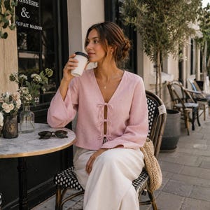 Puede incluir: Una blusa de color rosa claro de manga larga con escote en V y tres cierres de lazo en la parte delantera, combinada con pantalones blancos holgados. La blusa tiene un acabado de tela texturizada y mangas anchas, creando una silueta relajada.