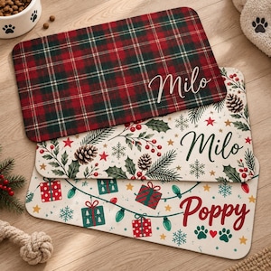 May include: Three rectangular pet mats with rounded corners. The top mat features a red and green plaid pattern with the name Milo. The middle mat has a white background with pine branches, pinecones, and berries, labeled Milo. The bottom mat displays colorful gift boxes, string lights, and paw prints with the name Poppy.